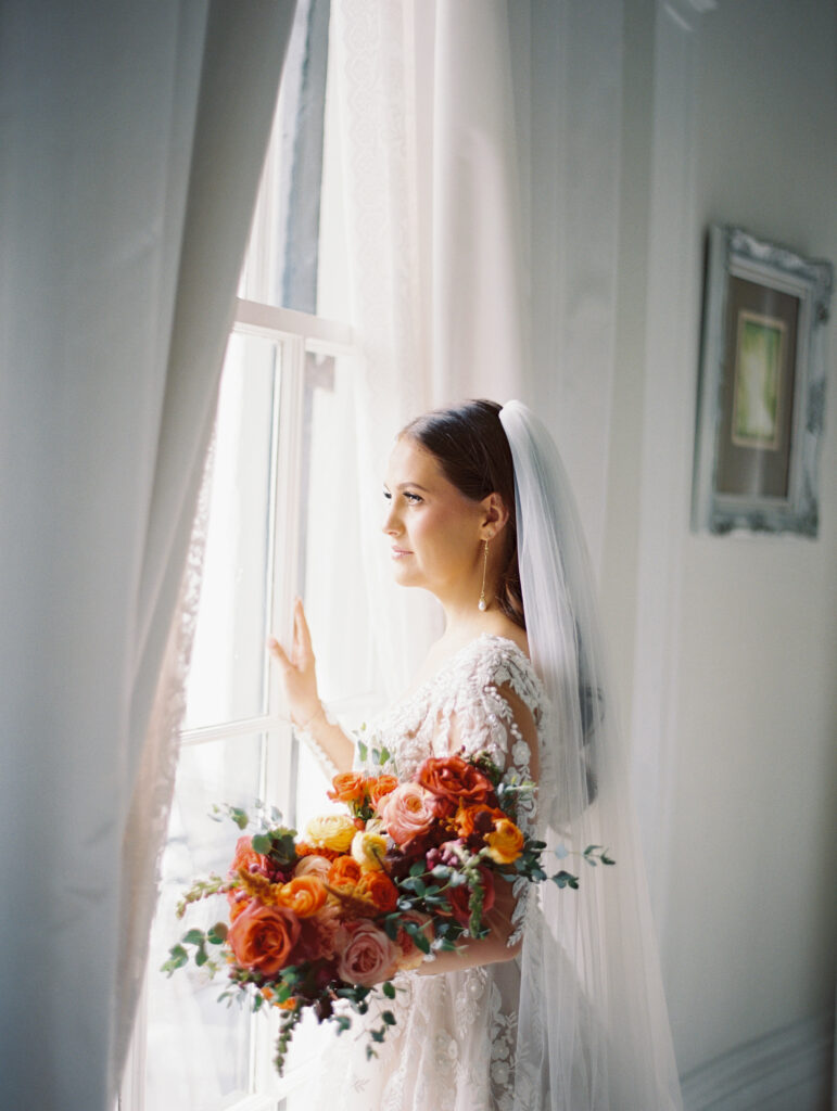 Close-up of a bride in a lace gown and veil holding a colorful bouquet, gazing out of a window with soft natural light. Photographed on film by Lexie Vaccaro Photography.