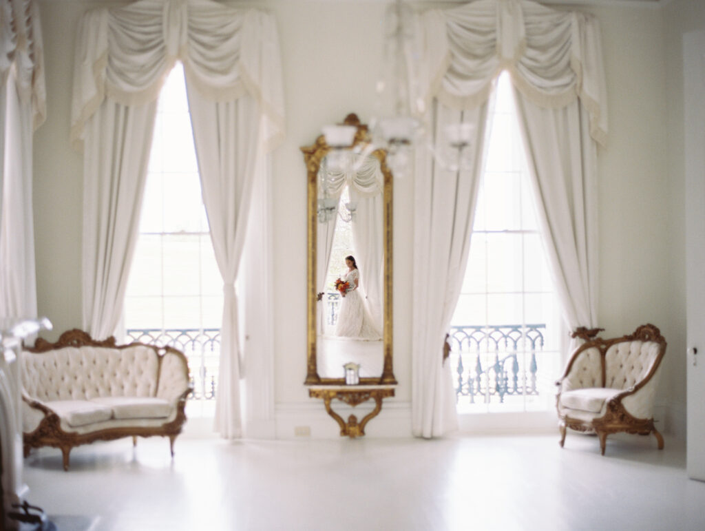 Reflection of a bride in a lace gown and veil holding a bouquet, captured in a gold-framed mirror within an elegant white room with vintage furniture. Photographed on film by Lexie Vaccaro Photography.