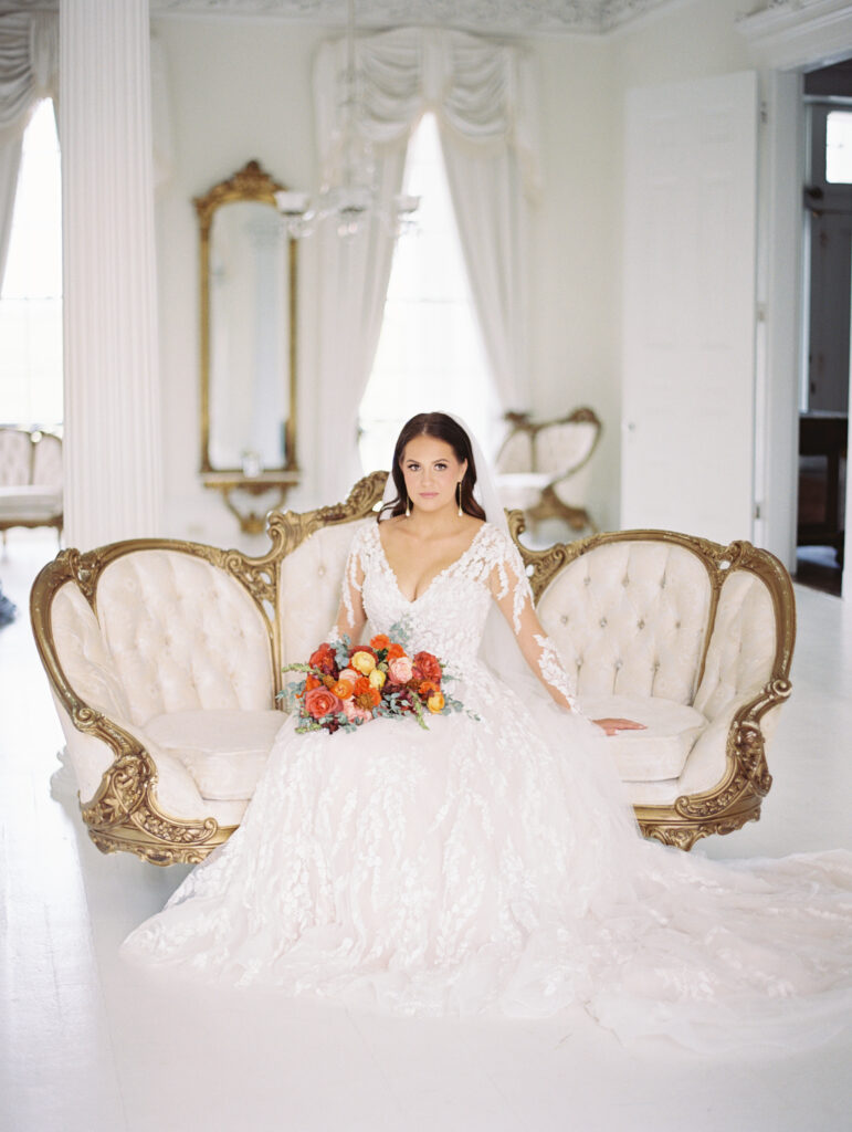 Bride in a lace gown and veil seated on a vintage gold-accented sofa, holding a vibrant bouquet in an elegant white room. Photographed on film by Lexie Vaccaro Photography.