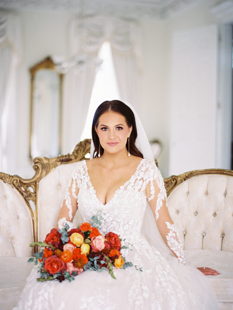 Bride in a lace gown and veil seated on a vintage tufted sofa with a vibrant bouquet, gazing confidently in an elegant white room. Photographed on film by Lexie Vaccaro Photography.