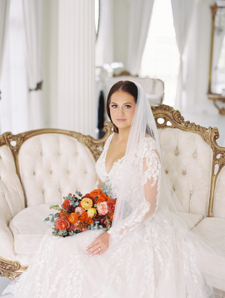 Bride in a lace gown and veil seated on a vintage gold-accented sofa, holding a vibrant bouquet in an elegant white room. Photographed on film by Lexie Vaccaro Photography.