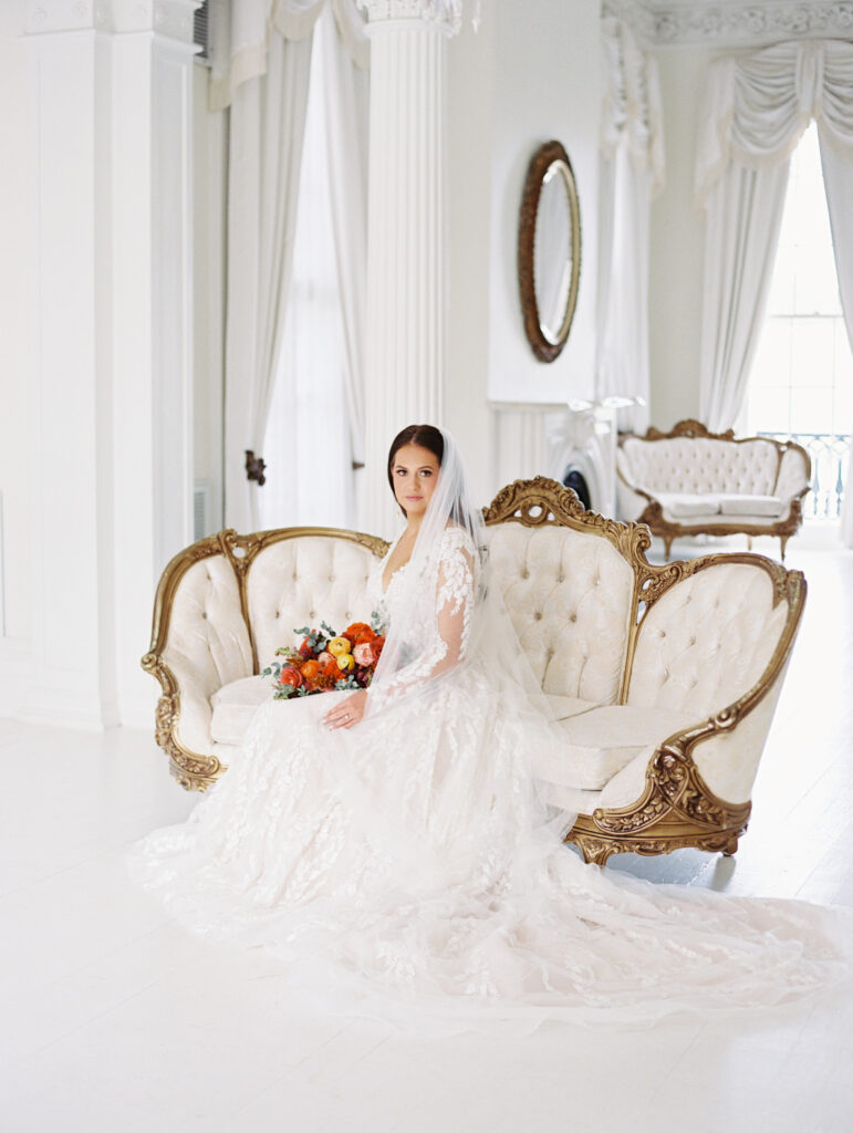 Bride in a lace gown and veil seated on a vintage tufted sofa with gold accents, holding a vibrant bouquet in an elegant white room. Photographed on film by Lexie Vaccaro Photography.