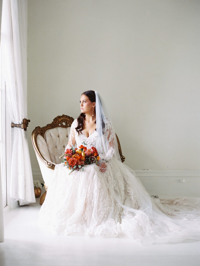 Bride in a lace gown and veil seated on a vintage chair, holding a vibrant bouquet, gazing out of a window in an elegant white room. Photographed on film by Lexie Vaccaro Photography.