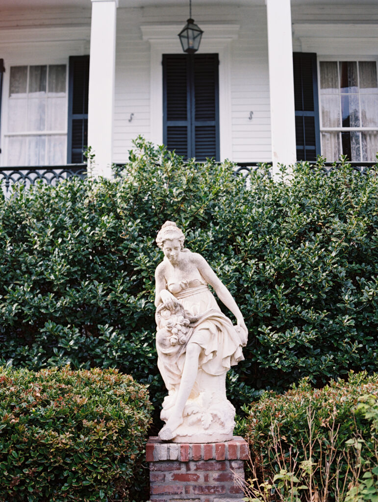 Stone statue of a woman with a floral arrangement surrounded by lush greenery, set against the classic Nottoway Plantation in Louisiana with shutters and a lantern. Photographed on film by Lexie Vaccaro Photography