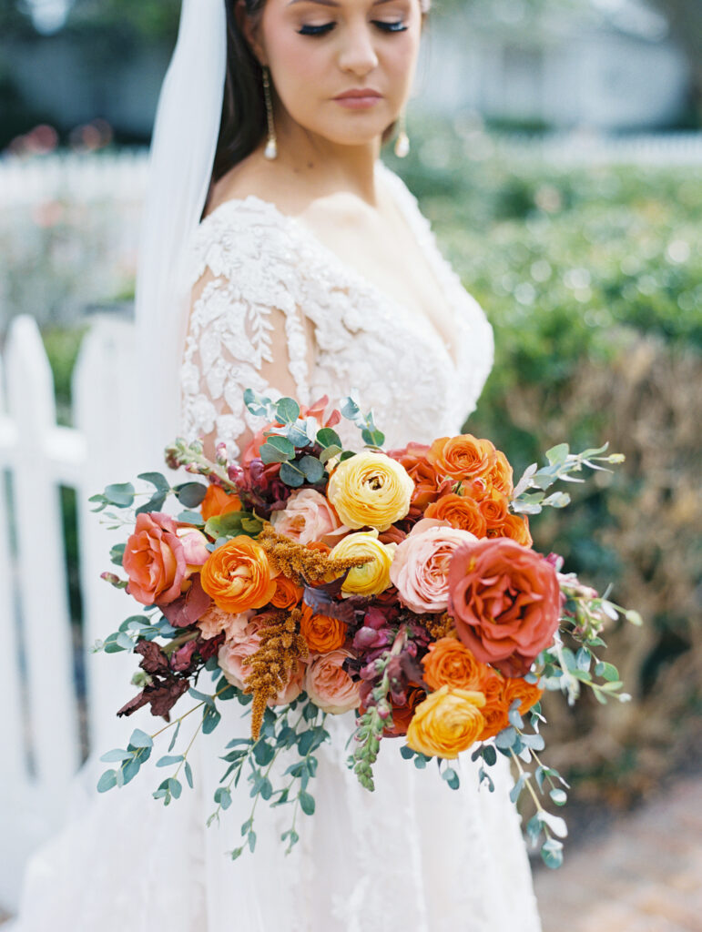 Close-up of a bride holding a vibrant bouquet with orange, yellow, and pink blooms, wearing a lace gown and veil in a garden setting. Photographed on film by Lexie Vaccaro Photography.