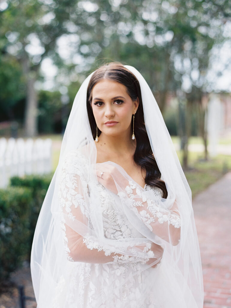 Bride in a lace gown and veil with delicate floral details, standing outdoors with a serene expression and soft natural light. Photographed on film by Lexie Vaccaro Photography.