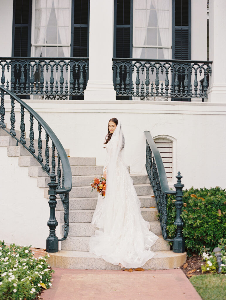 Bride in a lace gown and veil holding a vibrant bouquet, standing on elegant curved steps with a classic Southern balcony in the background at Nottoway in Louisiana. Photographed on film by Lexie Vaccaro Photography.