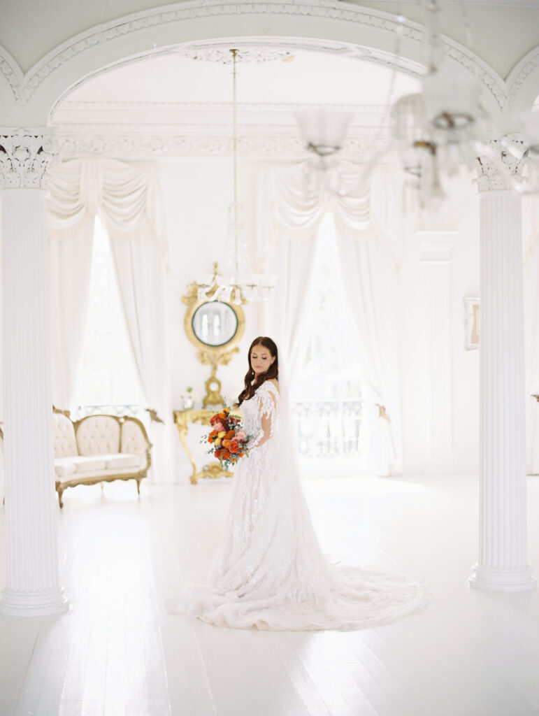 Bride in a lace gown and veil holding a vibrant bouquet, standing in an elegant white room framed by ornate columns and vintage decor. Photographed on film by Lexie Vaccaro Photography.