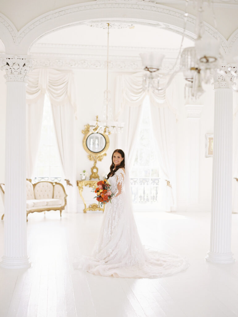 Bride in a lace gown and veil holding a vibrant bouquet, standing in an elegant white room framed by ornate columns and vintage decor. Photographed on film by Lexie Vaccaro Photography. At Nottoway in Louisiana.