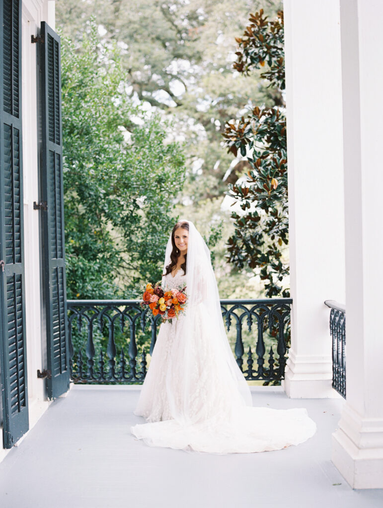 Bride in a lace gown and veil holding a vibrant bouquet, standing on a Southern-style balcony with lush greenery and classic shutters. Photographed on film by Lexie Vaccaro Photography.