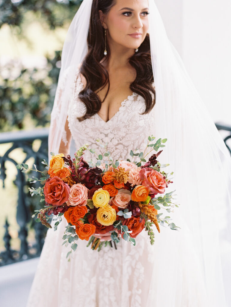 Bride in a lace gown and veil holding a vibrant bouquet of orange, yellow, and pink blooms, standing outdoors with soft natural light. Photographed on film by Lexie Vaccaro Photography.