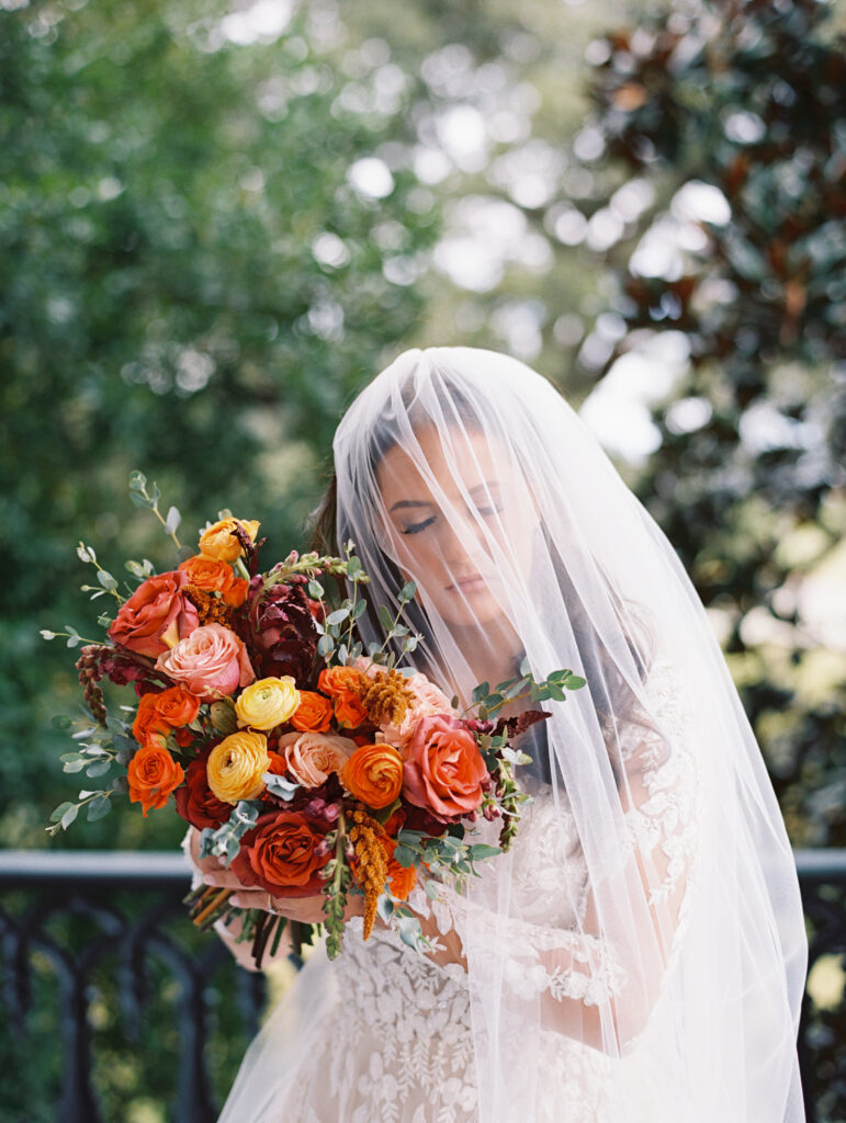 Bride in a lace gown and veil holding a vibrant bouquet of orange and yellow blooms, standing outdoors with greenery in the background. Photographed on film by Lexie Vaccaro Photography.