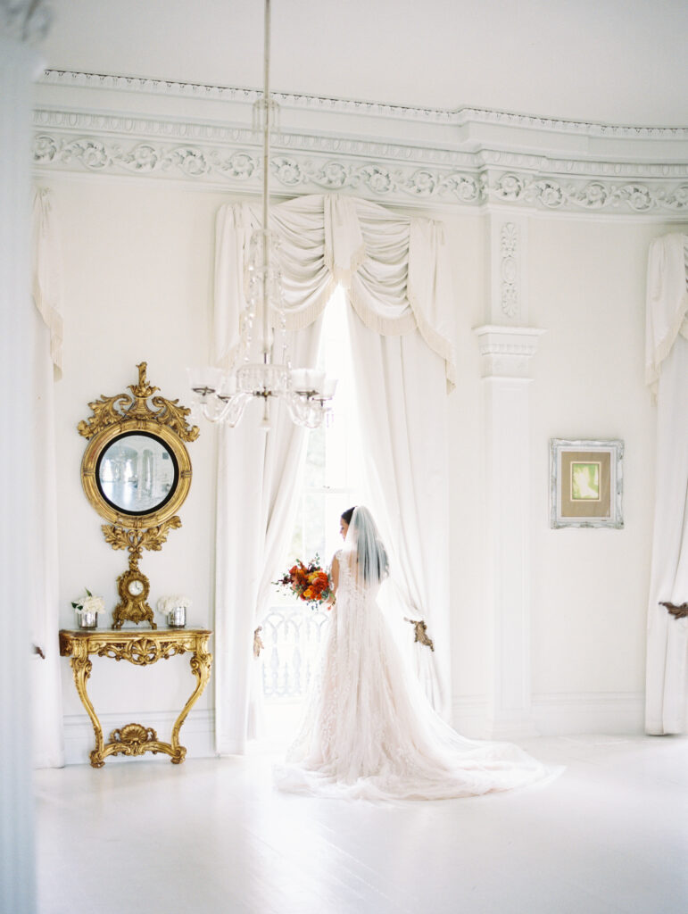 Elegant bride in a lace gown holding a vibrant bouquet, standing by a window in a luxurious white room with ornate gold details. Photographed on film by Lexie Vaccaro Photography.