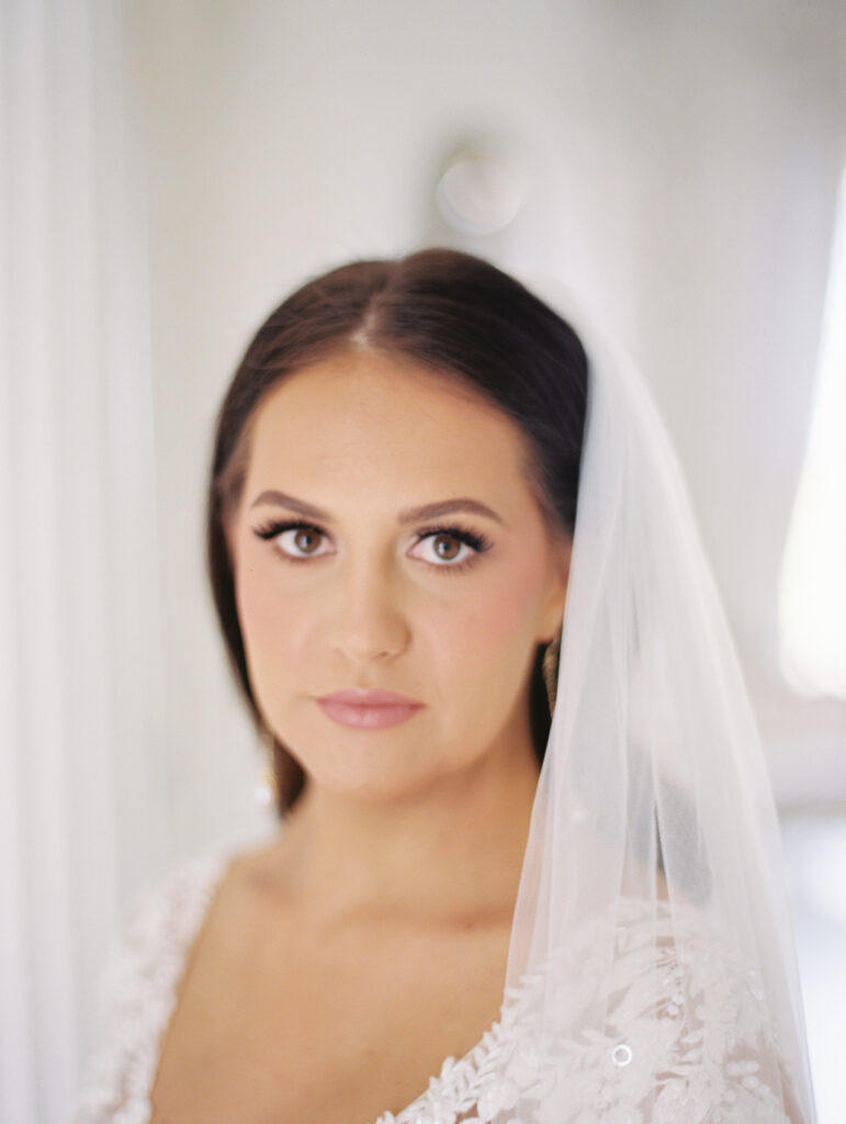 Close-up of a bride with a soft expression, wearing a lace gown and veil, captured in elegant natural light. Photographed on film by Lexie Vaccaro Photography.