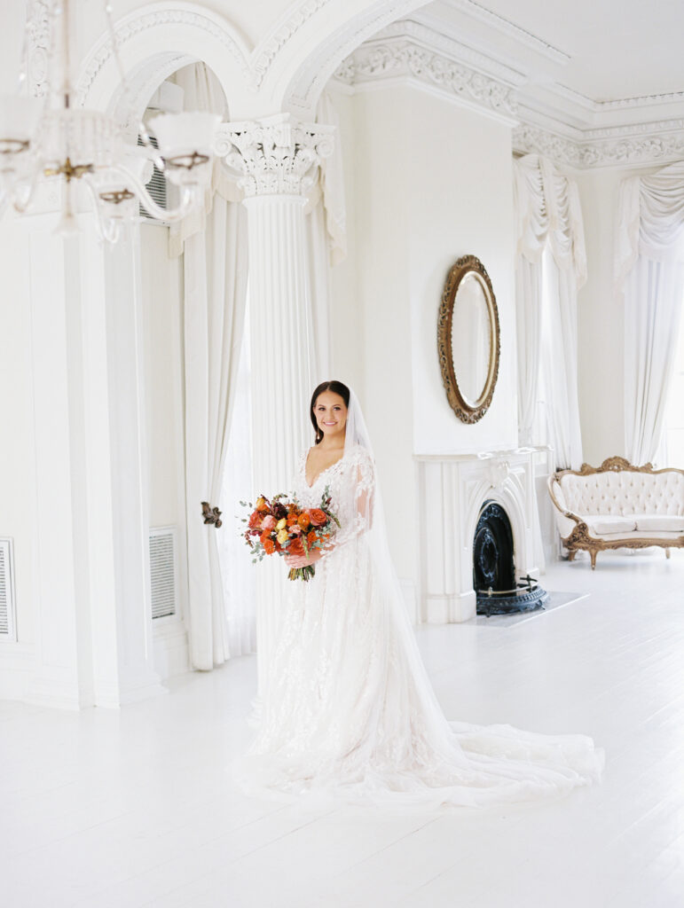 Bride in a lace gown and veil holding a vibrant bouquet, standing in an elegant white room with ornate columns and vintage decor. Photographed on film by Lexie Vaccaro Photography.