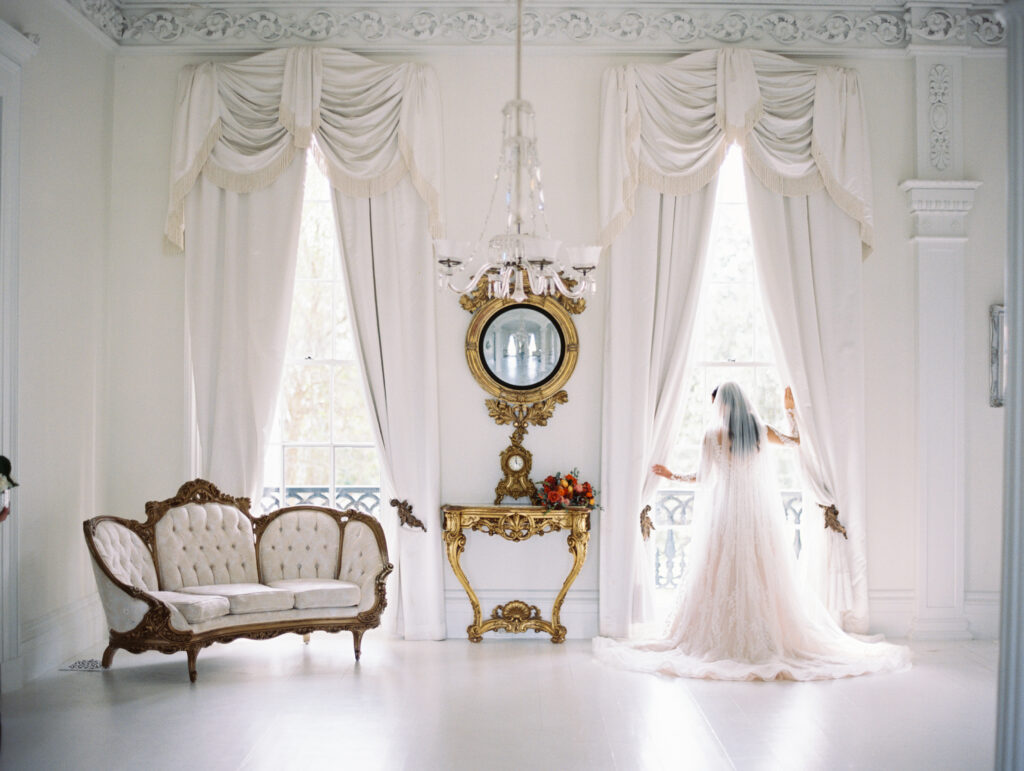 Bride in a lace gown and veil standing by a window in an elegant white room with vintage gold accents and a tufted sofa. Photographed on film by Lexie Vaccaro Photography.