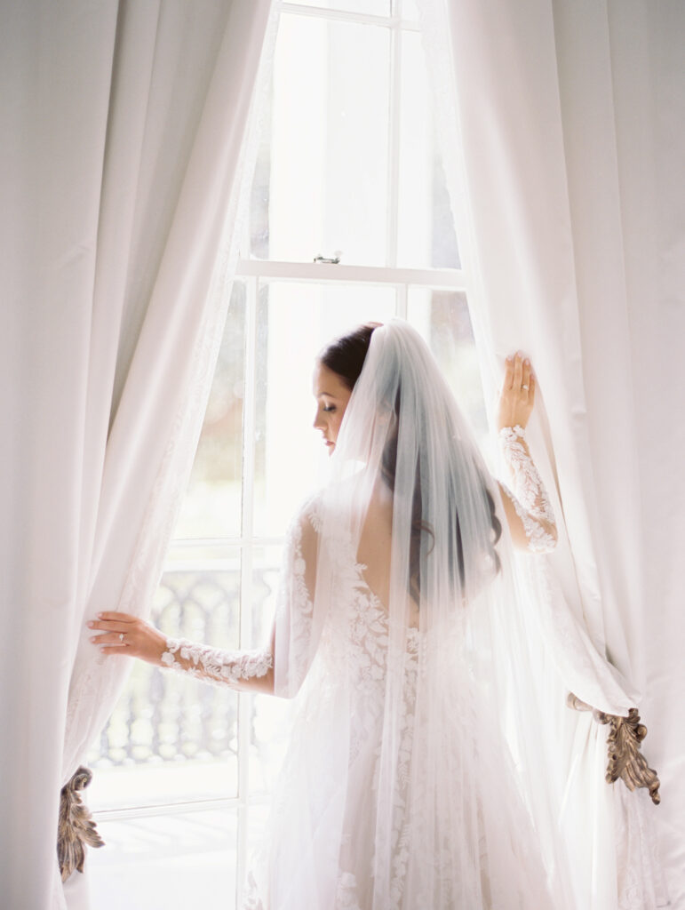 Bride in a lace gown and veil standing by a window, softly holding back the curtains in an elegant white room. Photographed on film by Lexie Vaccaro Photography