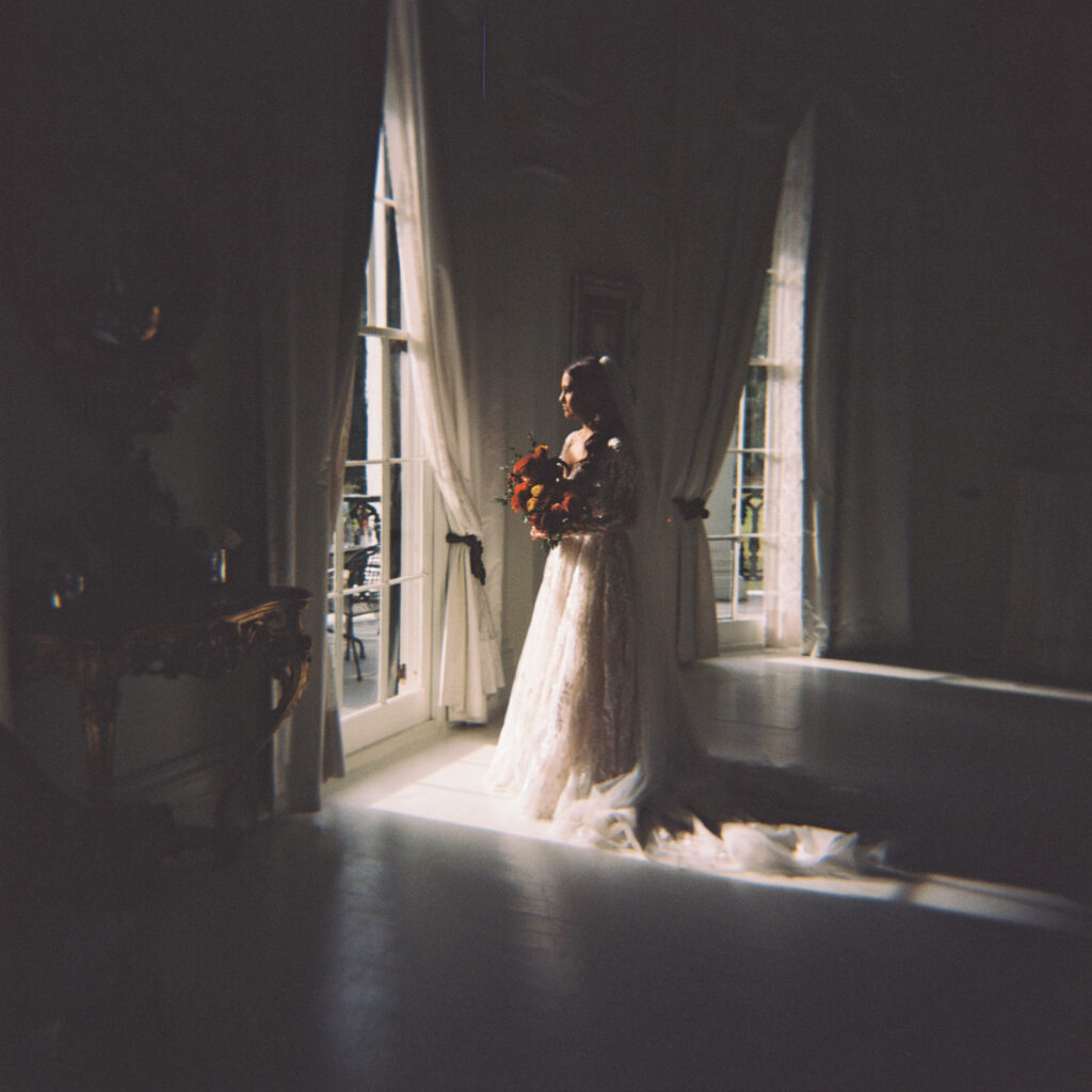 Moody bridal portrait of a bride standing in soft window light, holding a vibrant bouquet in an elegant white room with dramatic shadows. Photographed on film by Lexie Vaccaro Photography.