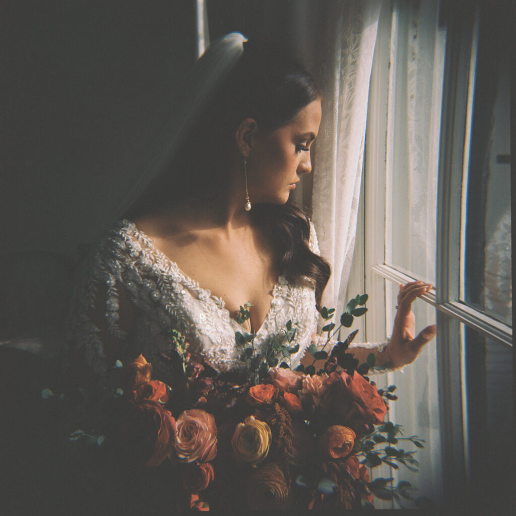 Moody bridal portrait of a bride in a lace gown and veil holding a vibrant bouquet, gazing out of a window with soft light and dramatic shadows. Photographed on film by Lexie Vaccaro Photography.