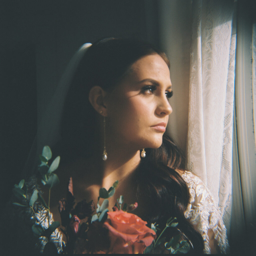 Moody bridal portrait with dramatic light highlighting the bride's serene profile, wearing a lace gown and holding a bouquet with vibrant blooms. Photographed on film by Lexie Vaccaro Photography.