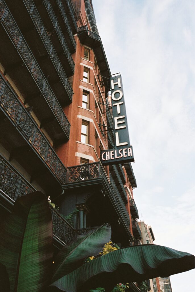 Historic Hotel Chelsea in New York City with its iconic black wrought iron balconies and vertical neon sign against a blue sky, framed by lush green leaves.