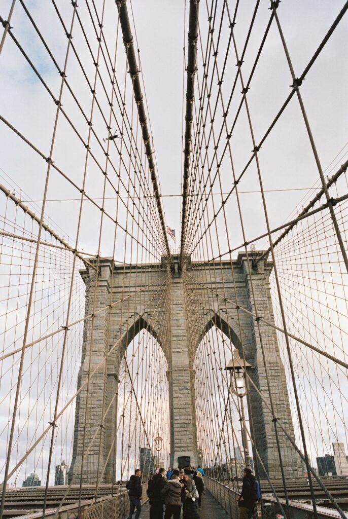 Film photo of the Brooklyn Bridge cables and arches, captured with people walking along the pathway at sunset, taken on Kodak Portra 400 film with a Contax G1 camera.