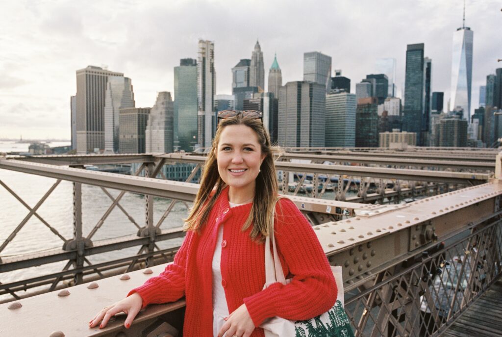 Portrait of Lexie Vaccaro standing on the Brooklyn Bridge in a red sweater with the Manhattan skyline in the background, captured on Kodak Portra 400 film.