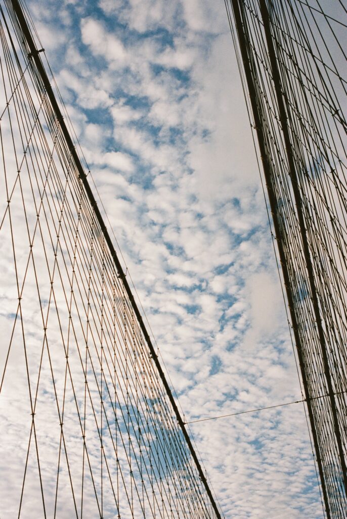 Film photo of the Brooklyn Bridge cables stretching upward against a blue sky with scattered clouds, captured on Kodak Portra 400 film with a Contax G1 camera.