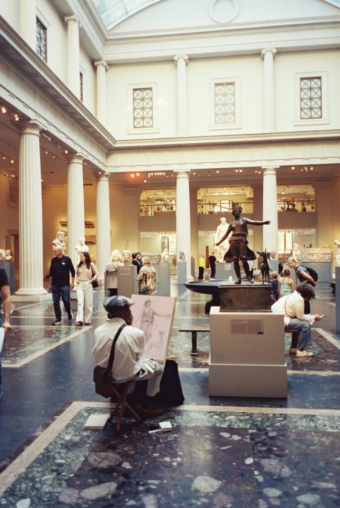 An artist sketching a statue inside the Metropolitan Museum of Art, surrounded by visitors admiring sculptures and exhibits in a grand hall with columns and high ceilings.