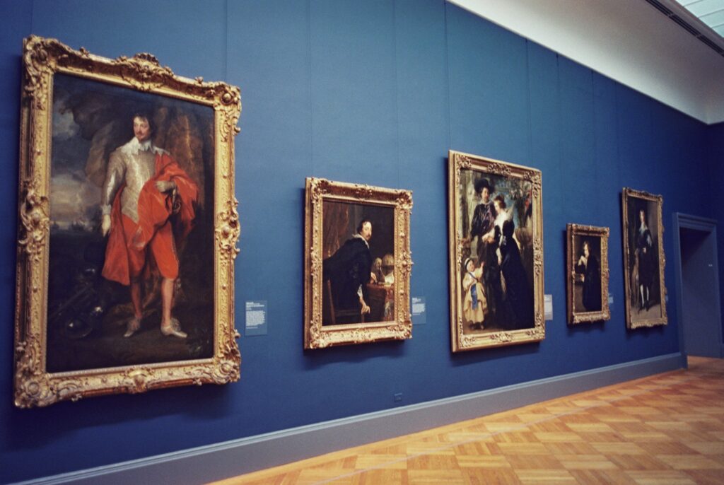 A gallery inside the Metropolitan Museum of Art featuring an array of framed portraits displayed on a deep blue wall, with ornate gold frames and a polished wooden floor.