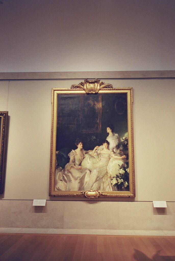 A portrait painting displayed at the Metropolitan Museum of Art, showcasing three women elegantly dressed in white gowns, seated and posed in an ornate setting.