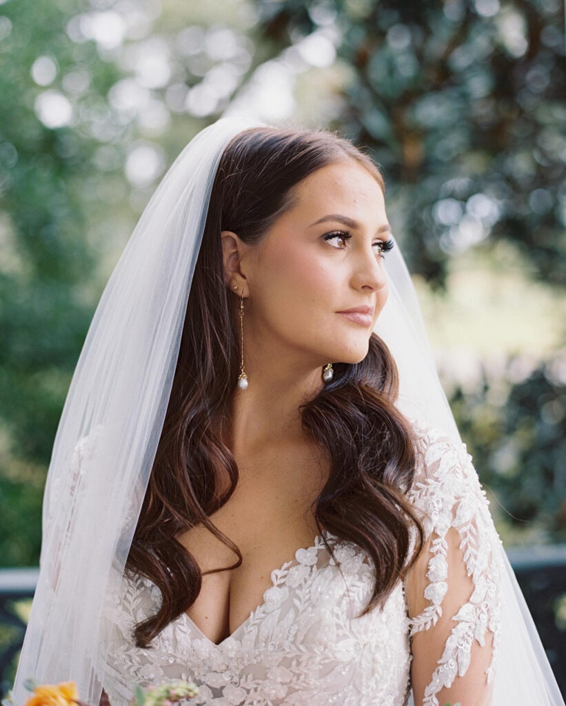 Close-up of a bride in a lace gown and veil with soft curls, gazing to the side against a backdrop of greenery. Photographed on film by Lexie Vaccaro Photography.