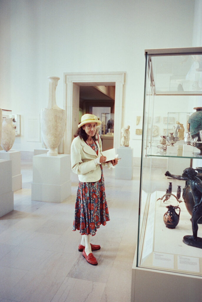 Film photo of an artist sketching in a gallery at The Metropolitan Museum of Art, wearing a colorful floral dress and yellow hat, surrounded by ancient artifacts, captured on CineStill film with a Contax G1 camera.