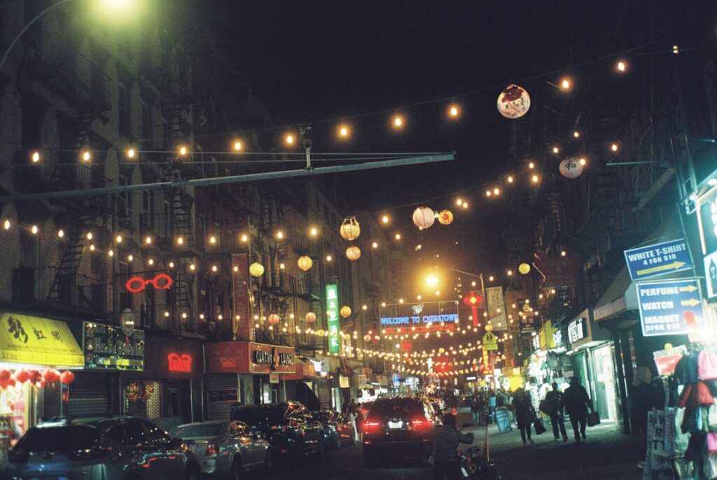 Chinatown at night in New York City, illuminated with strings of glowing lanterns and colorful neon signs, creating a lively and atmospheric street scene.