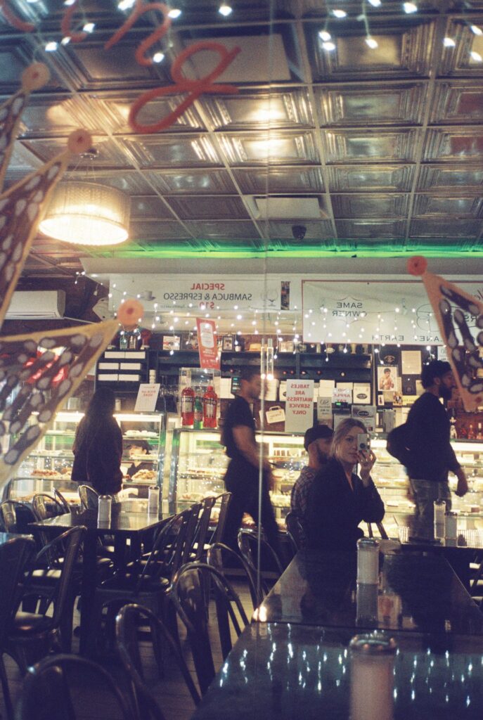 Interior of a lively cafe at night, with reflective metallic ceilings, a counter filled with desserts, and a soft glow of string lights. A seated woman captures a selfie amidst the bustling atmosphere.