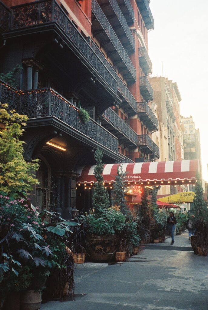 Historic Hotel Chelsea facade with intricate black wrought iron balconies and a red and white striped awning, surrounded by lush greenery, captured on Cinestill film.