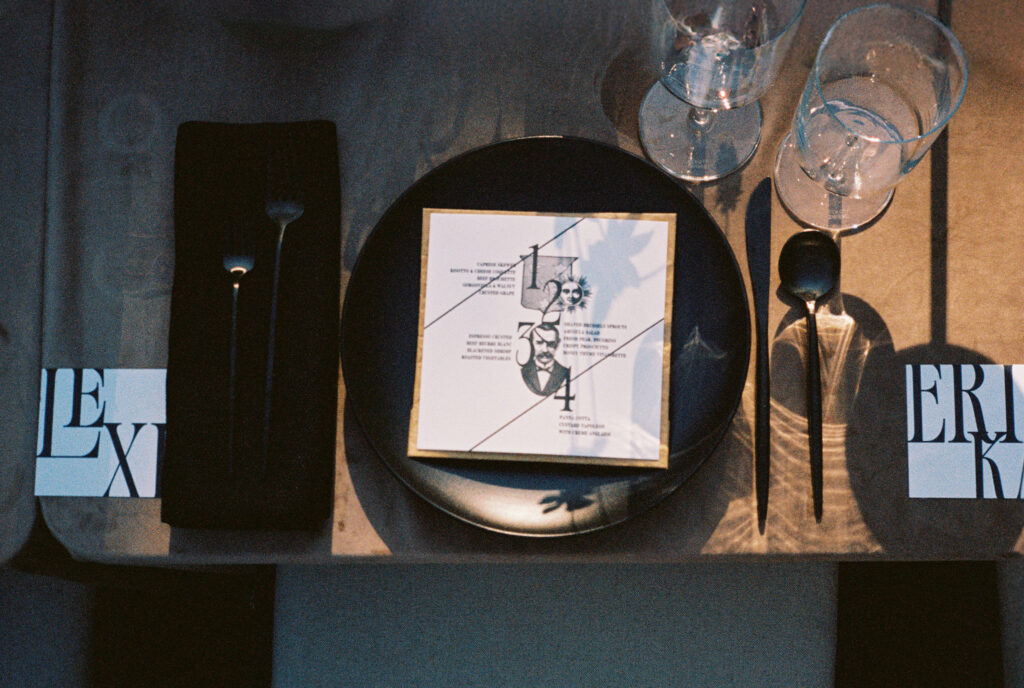 A moody, artistic place setting at Greencrest Manor, featuring black dinnerware, modern typography menus, and delicate shadows cast by glassware and florals.