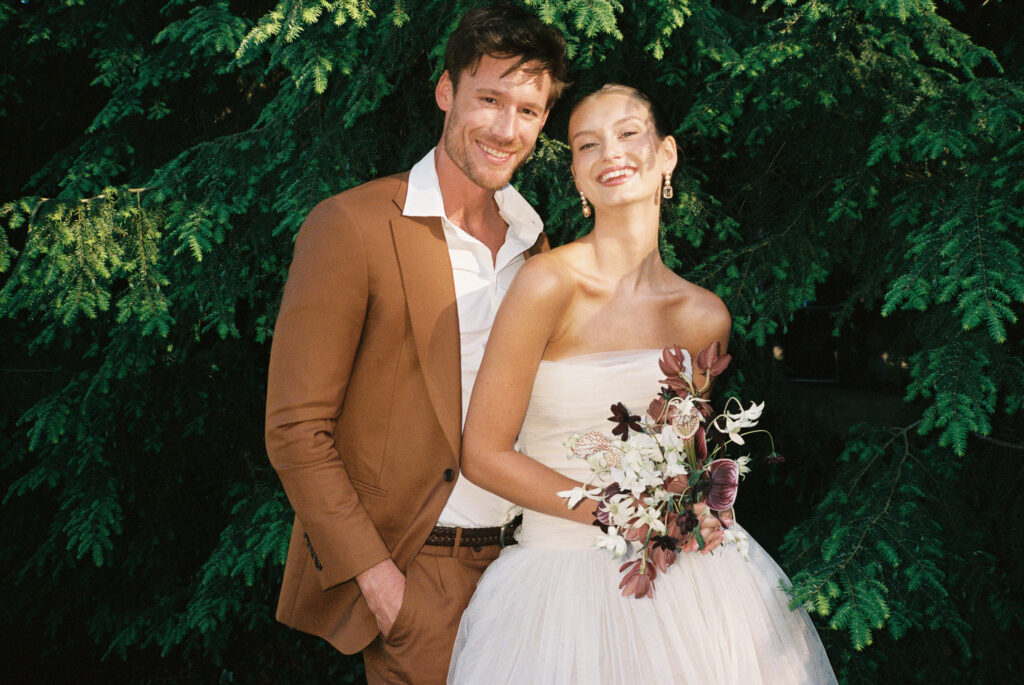 A joyful bride and groom pose against lush greenery at Greencrest Manor, captured on film by Lexie Vaccaro Photography. The bride wears a strapless gown with a modern bouquet, while the groom dons a camel-hued suit.