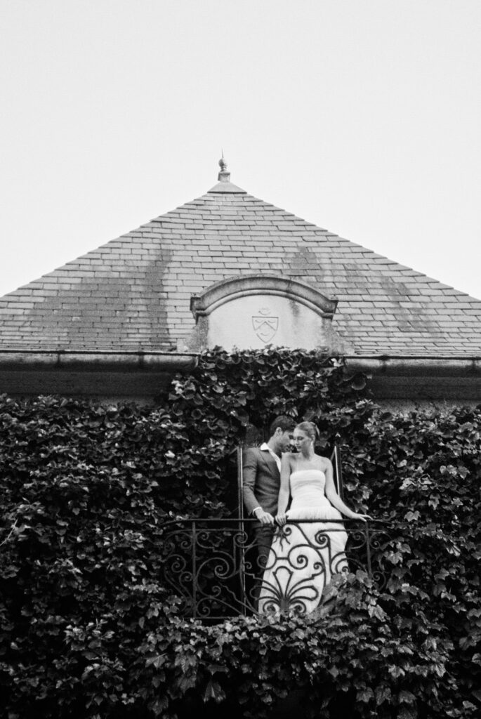 A black-and-white film portrait of a bride and groom embracing on a wrought-iron balcony covered in ivy at Greencrest Manor, evoking a timeless, cinematic romance.