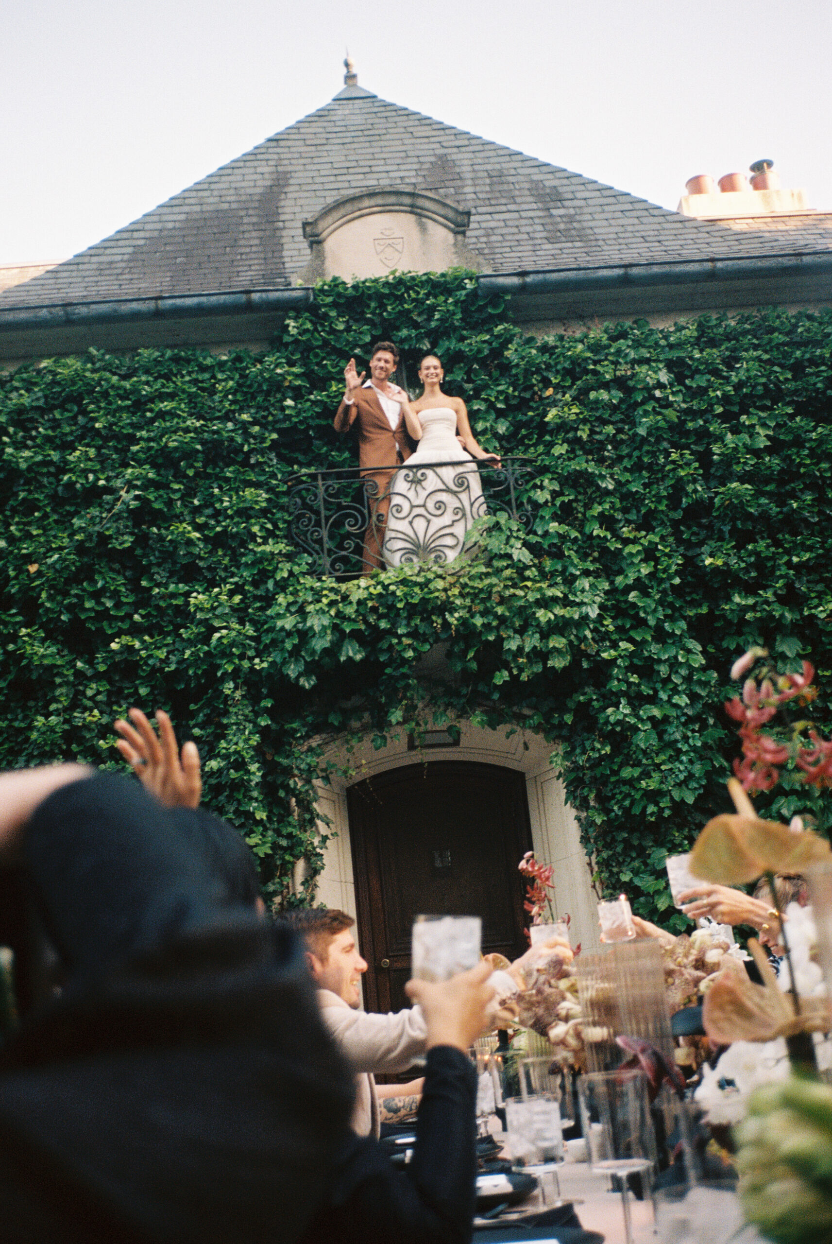 Bride and groom wave from a romantic ivy-clad balcony at Greencrest Manor as wedding guests raise their glasses in a celebratory toast.