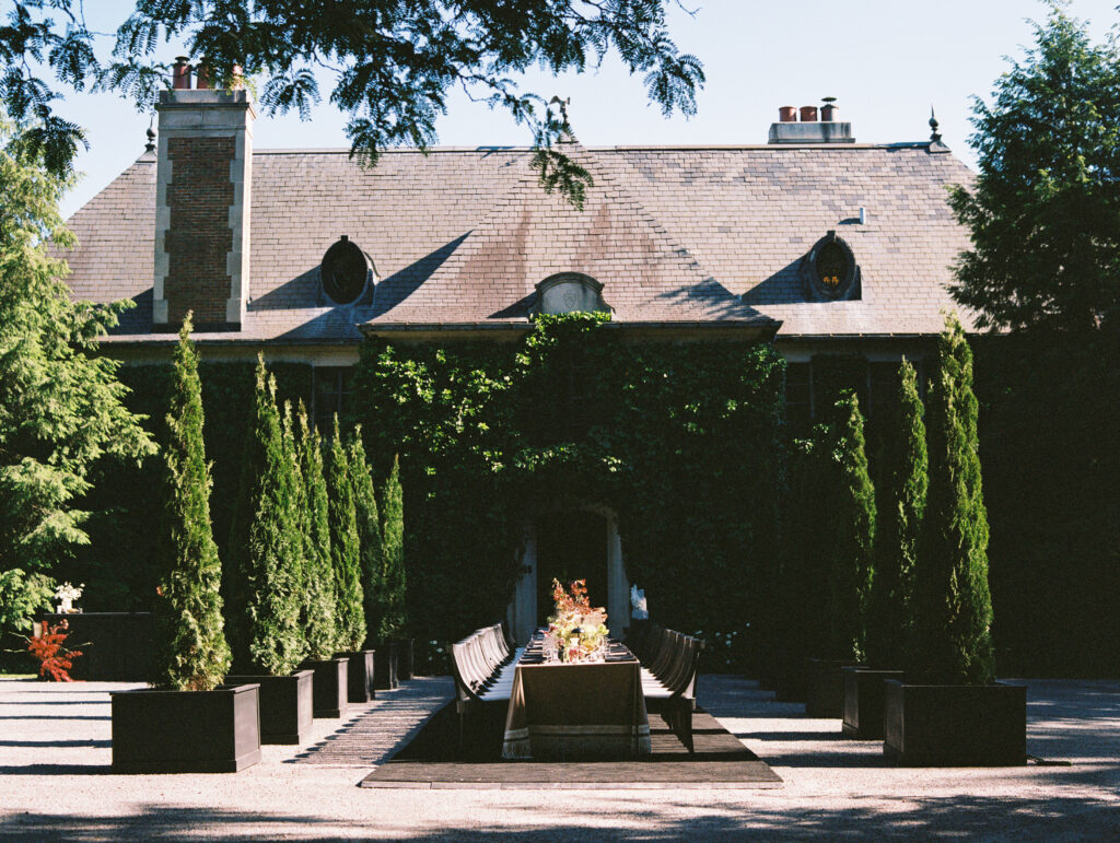 A long, elegant wedding reception table set in front of the ivy-covered Greencrest Manor, framed by towering cypress trees and European-inspired architecture.