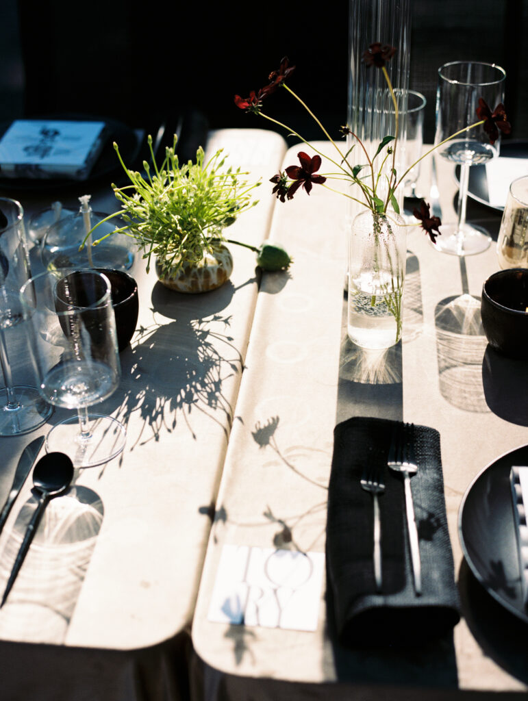 A modern wedding tablescape with minimalist floral arrangements, black tableware, and soft golden light casting artistic shadows on the table, captured on film.