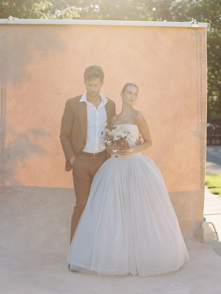 A bride in a strapless Lein Studio gown and groom in a brown suit pose against a warm-toned backdrop, bathed in golden film light for an ethereal, editorial wedding portrait.