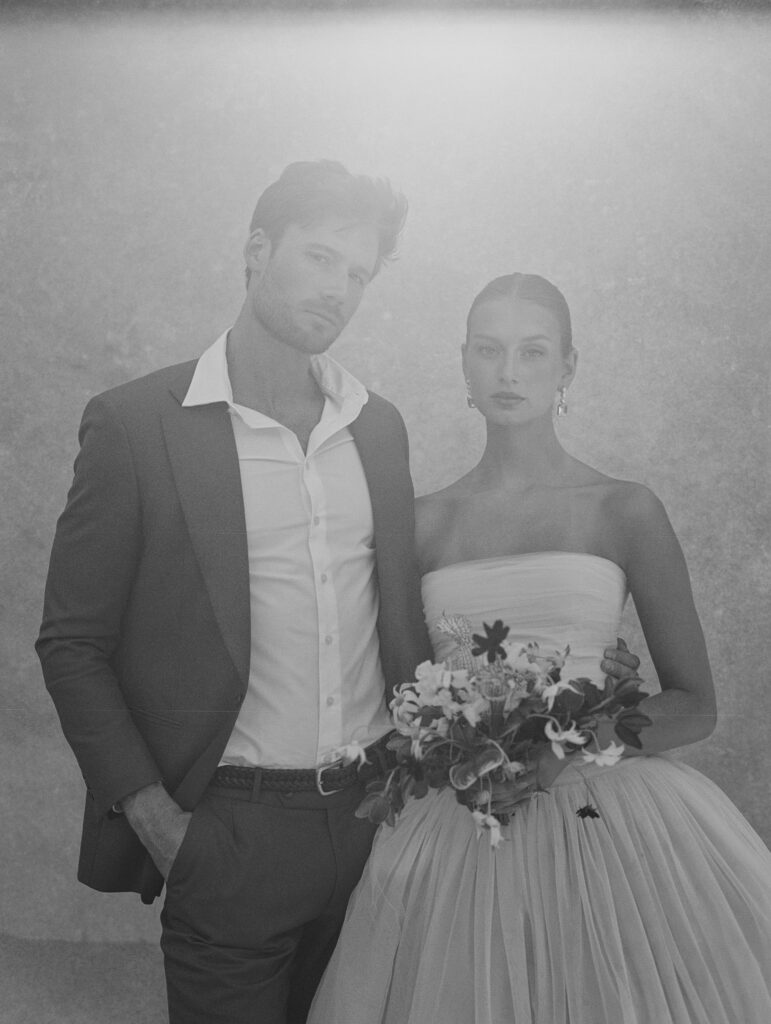 A black-and-white film portrait of a bride in a strapless Lein Studio gown and groom in a tailored suit, standing against a textured Chasing Stone backdrop with soft, ethereal light.