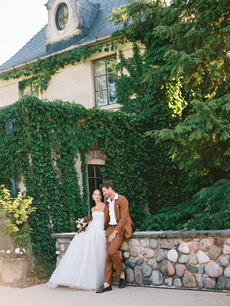 A bride and groom share a relaxed, romantic moment outside the ivy-covered Greencrest Manor, bathed in warm afternoon light. Captured on film for an editorial, old-world aesthetic.