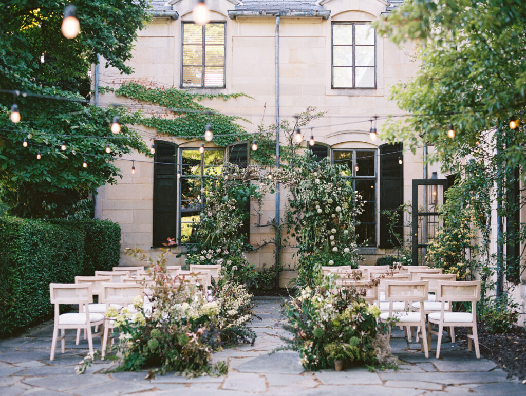 A romantic outdoor wedding ceremony at Greencrest Manor, featuring lush floral installations, natural wood chairs, and string lights draped across the courtyard.