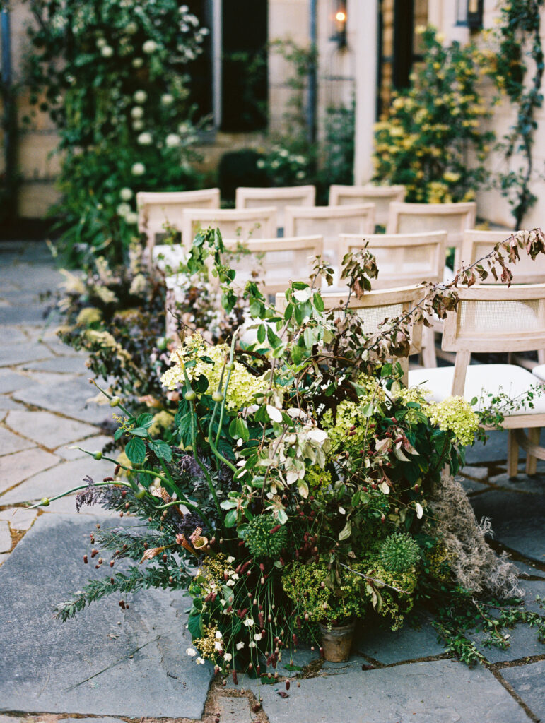 Lush, organic floral arrangements line the aisle of a European-inspired wedding ceremony, featuring delicate blooms and textured greenery.