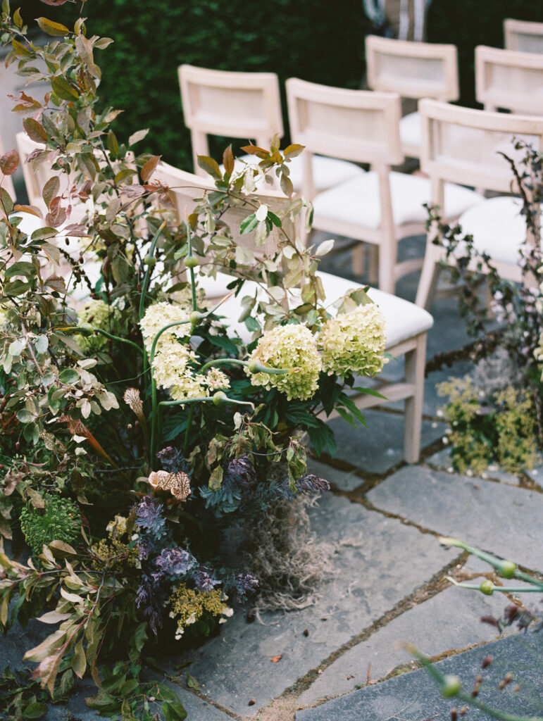 A close-up of an organic floral arrangement with lush hydrangeas and wild greenery lining the ceremony aisle, with natural wood chairs set on a stone pathway at Greencrest Manor.