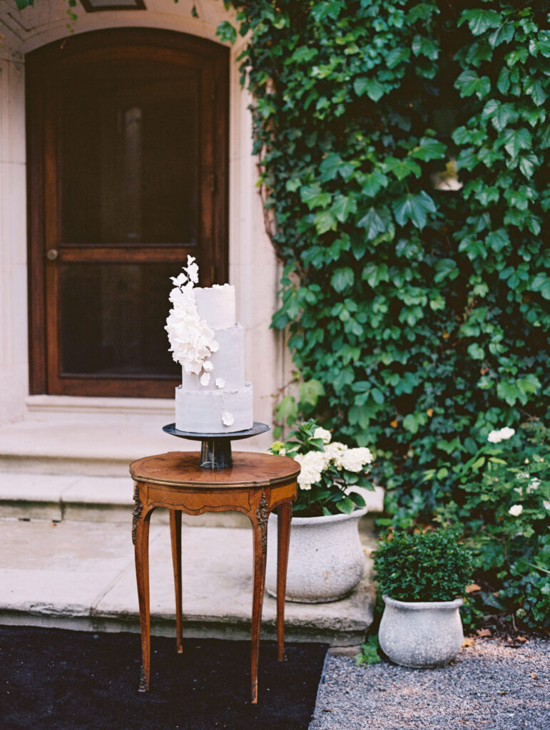 A modern, sculptural wedding cake with delicate sugar flowers sits atop an antique wooden table, framed by lush greenery and an ivy-covered stone doorway.