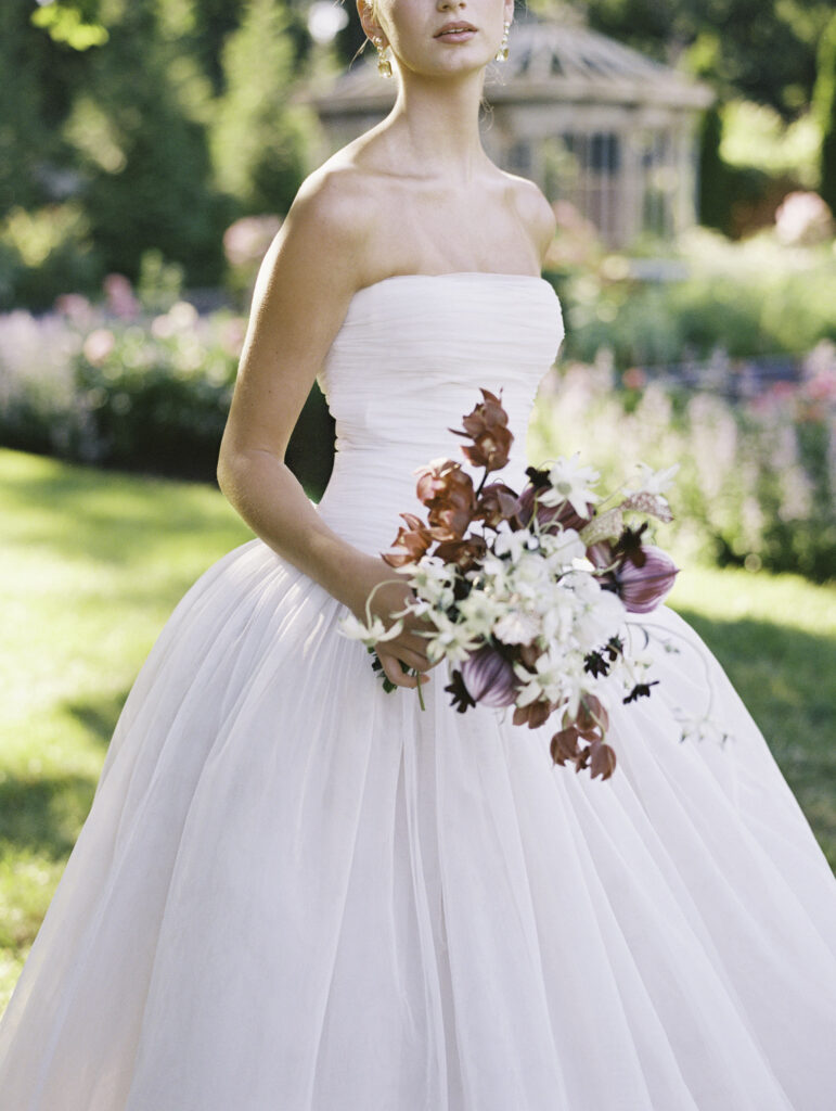 A bride in a strapless Lein Studio gown holds a sculptural bouquet in the gardens of Greencrest Manor, with a vintage conservatory softly blurred in the background, captured on film.